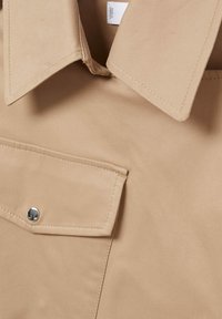 Gros plan du col de chemise beige et de la poche poitrine avec bouton-pression argenté, montrant la texture du tissu et les détails de couture.