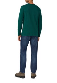 Mann in dunkelgrünem Langarmhemd, blauen Jeans und braunen Schuhen, der mit dem Rücken zu einem weißen Hintergrund steht.