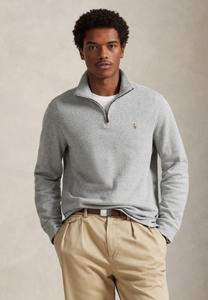 Homme portant un pull gris clair à fermeture éclair quart de zip sur une chemise blanche et un pantalon beige, debout les mains dans les poches devant un fond uni.