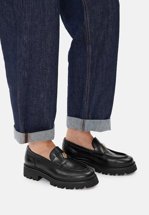 Mocassini neri in pelle con suola spessa e dettagli in oro, abbinati a pantaloni di denim scuri arrotolati con risvolto azzurro chiaro.