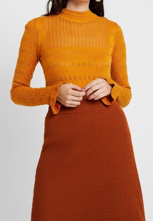 Frau trägt einen strukturierten orangefarbenen Strickpullover mit gerafften Bündchen und einem rostfarbenen, gerippten Strickrock, Hände sanft an der Taille verschränkt.