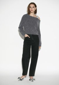 Grijze off-shoulder sweater, high-waisted zwarte jeans met verfspatten en zilveren hakken met strikdetails.