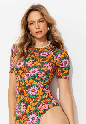 Femme aux longs cheveux blonds portant un body à manches courtes fleuri et coloré et un collier de perles blanches sur un fond uni.