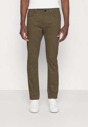 Hombre con pantalones ajustados verde oliva con bolsillos delanteros y zapatillas blancas, de pie contra un fondo blanco liso.