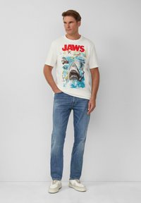 Weißes T-Shirt mit einem roten "JAWS"-Grafikdesign, das einen Hai und eine Wasserszene zeigt. Kombiniert mit blauen Jeans und weißen Sneakers.