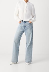 Kvinde iført en hvid langærmet bluse med volangdetaljer, stoppet ned i lyseblå vide jeans og hvide spidse hæle, hænderne i lommerne.