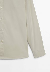 Jasnobeżowa koszula zapinana na guziki o gładkiej fakturze, z długimi rękawami i lekko zaokrąglonym dołem. Posiada pięć guzików i klasyczny krój kołnierzyka.