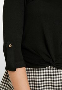 Svart, räfflad stickad topp med rullade ärmar och en accent av en träknapp, knuten framdesign. Bärs över en svart och vit rutmönstrad kjol.