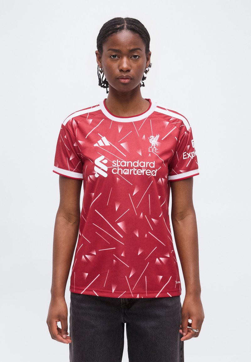 Γυναίκα που φοράει κόκκινη φανέλα της Liverpool FC με λευκά γεωμετρικά σχέδια και λογότυπα χορηγών, που στέκεται μπροστά σε λευκό απλό φόντο.