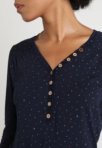 Kvinna som bär en marinblå långärmad skjorta med små stjärnmönster och en rad träknappar vid V-ringningen.