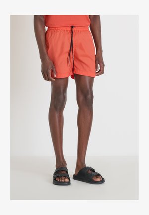 Bañador naranja brillante con cintura elástica y cordón negro. Combinado con sandalias negras sin hebilla. Tejido suave y ligero.