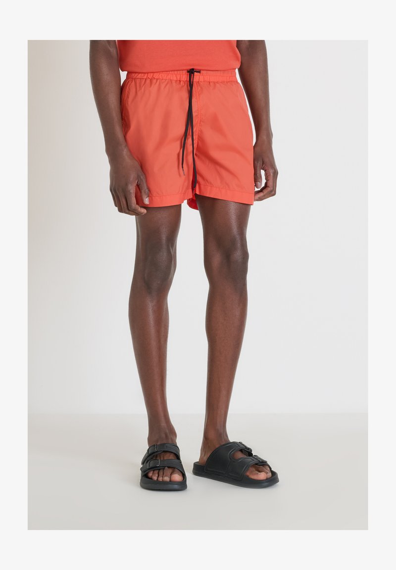 Bañador naranja brillante con cintura elástica y cordón negro. Combinado con sandalias negras sin hebilla. Tejido suave y ligero.