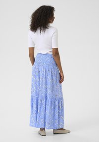 Jupe maxi en bleu clair avec un motif ondulé, taille smockée et volants en tiers. Associée à un haut ajusté blanc et des ballerines beiges.