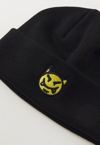 Must have mustrikoot punane beanie, millel on kollane tikandiga naeratav nägu, millel on sarved ja hambad. Pehme tekstuur ja rullitud varrukate disain.