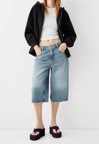 Culottes de mezclilla en azul claro con un corte holgado, dobladillo deshilachado y bolsillos delanteros, combinados con una sudadera con capucha negra con cremallera y sandalias con plataforma.