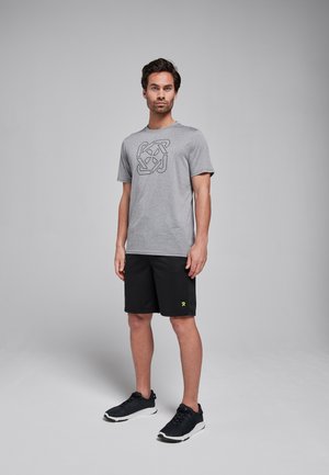 Grijze t-shirt met korte mouwen en een zwart geometrisch patroon, gecombineerd met zwarte shorts en zwarte sportieve schoenen met een gele logo.