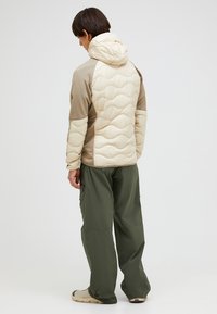 Giacca isolata beige e marrone con cappuccio, caratterizzata da un motivo a zigzag trapuntato. Indossata con pantaloni cargo verde scuro e scarpe chiare.
