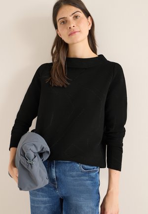 Suéter negro texturizado con cuello redondo, mangas largas y patrones en relieve sutiles, combinado con jeans de mezclilla azul y una chaqueta gris.
