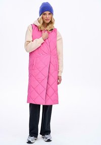 Różowa pikowana długa kamizelka z zapięciem na zatrzaski z przodu, kieszeniami bocznymi i wysokim kołnierzem, noszona na beżowym hoodie i czarnych spodniach.