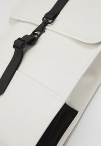 Mochila blanca con un acabado suave, que cuenta con una correa negra y herrajes negros. Incluye una solapa superior y un bolsillo lateral con forro de tela negra.