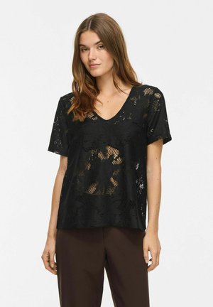 Femme portant un top en dentelle noire à manches courtes avec un décolleté en V et un pantalon marron, debout devant un fond blanc uni.