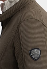 Brun mönstrad jacka med hög krage, silverdragkedja och en svart sköldlogotyp på vänster ärm, med en texturerad yta.