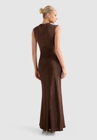 Sieviete ar blondiem matiem sasietiem bulciņā, valkājot bezroku, piegulošu, līdz grīdai sniedzošu spīdīgu brūnu kleitu, stāvot un skaties sāniski pret vienkāršu fonu.