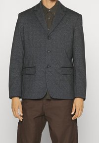 Mörkgrå blazer i mönstrad tyg, med enfaldsdesign, utskurna slag och två framfickor. Bärs över en grön skjorta.