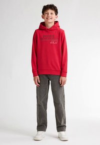 Červená bavlněná mikina s kapucí s šedým logem, v kombinaci se šedými manšestrovými kalhotami a bílými teniskami, stojící proti jednobarevnému pozadí.