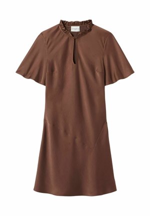 Femme debout portant une robe marron à manches courtes avec un pull beige sur les épaules, des chaussettes rouges et des chaussures marron à lacets.