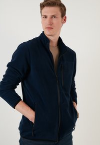 Navy blue fleece jacket with a high collar, two zippered front pockets, and textured cuffs. Features a sleek, fitted design.