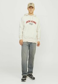 Sudadera de color claro con logo rojo, jeans azules desgastados y zapatillas negras con detalles blancos. La gorra es beige con una letra oscura.