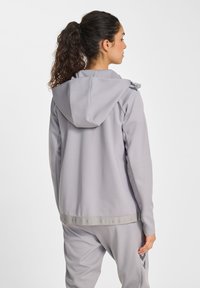 Ljusgrå sportjacka med huva, mjuk textur och diskret logodetalj vid fållen. Matchas med joggingbyxor i samma färg.