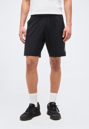 Άνδρας που φοράει μαύρα αθλητικά σορτς, λευκές κάλτσες, μαύρα αθλητικά παπούτσια Adidas και λευκό πουκάμισο, στεκόμενος μπροστά σε απλό ανοιχτόχρωμο φόντο.