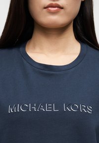 T-shirt de algodão azul-marinho com decote redondo, com texto branco em relevo "MICHAEL KORS" atravessando o peito. Textura lisa.