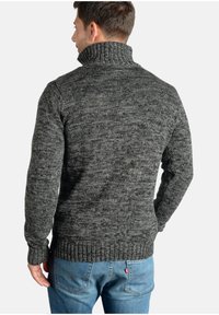 Jersey de cuello alto gris oscuro con un patrón texturizado y puños acanalados, combinado con jeans azules. El jersey presenta un corte holgado.