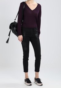 Pull à col en v violet foncé, jean skinny noir avec des détails déchirés, baskets noires et beiges, et un sac à bandoulière noir.