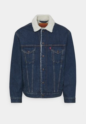 Dunkelblaue Jeansjacke mit weißem Sherpa-Kragen, Knopfleiste vorne und zwei Brusttaschen mit Knopflappen, dargestellt auf weißem Hintergrund.