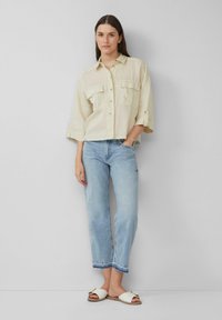 Helle beige Leinenbluse mit zwei Fronttaschen und gekürzten Ärmeln. Kombiniert mit hellblauen Jeans und weißen Riemchensandalen mit goldenen Akzenten.