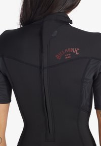 Čierny neoprénový oblek s krátkymi rukávmi, ktorý má textúrované vzory a vertikálny zips na zadnej strane. Obsahuje červený detail loga na hornej časti chrbta.