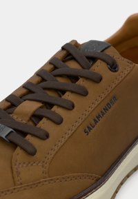 Sapatilha de couro castanho com atacadores castanhos escuros, detalhes cosidos e uma pequena etiqueta de logotipo preta. Sola de borracha branca com piso texturizado.