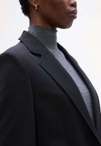 Černý blazer s ostrými klopami vyrobený z hladké látky, přes šedý rolák. Detailní záběr zaměřený na límec a texturu.
