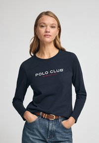 T-shirt a manica lunga navy con logo "POLO CLUB" in bianco e rosso, colletto tondo, indossato con jeans in denim e una cintura marrone, texture del tessuto liscia.