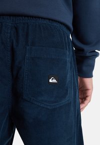 Navy corduroy broek met een elastische tailleband, voorzien van een achterzak en een klein logo-label. Verticale ribbelstructuur.