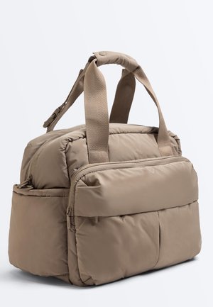 Béžová polstrovaná duffel taška so širokými textilnými rukoväťami, predným vreckom na zips, bočnými vreckami a odnímateľným ramenným popruhom na bielom pozadí.