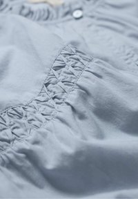 Hellblaues Gewebe mit detaillierter Smocking-Stickerei und Nahtnähten in der Nähe eines geknöpften Kragens, das die Textur und das textile Muster im Nahbereich zeigt.