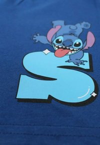 T-shirt en coton bleu avec un personnage de cartoon aux grandes oreilles et une langue rose, accompagné d'une lettre "S" bleue en gras. Texture lisse, design vibrant.