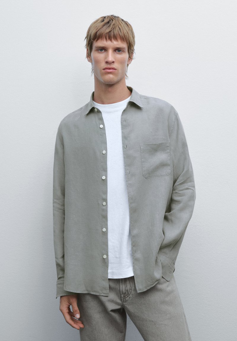 Giovane in camicia grigia chiaro con bottoni sopra una maglietta bianca e jeans grigi, in piedi contro una parete grigia chiaro uniforme.