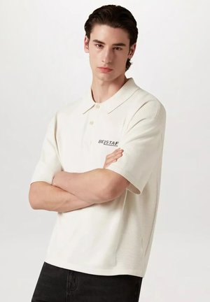 Junger Mann mit dunklem Haar, trägt ein offweißes BE|STAF Poloshirt, Arme verschränkt, vor einem schlichten hellen Hintergrund stehend.