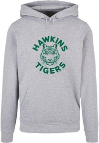Pelēks kapuci džemperis ar centrālo izdrukāto dizainu, uz kura redzams tigers un teksts "HAWKINS TIGERS" zaļā krāsā. Iekļauta priekšējā kabata.